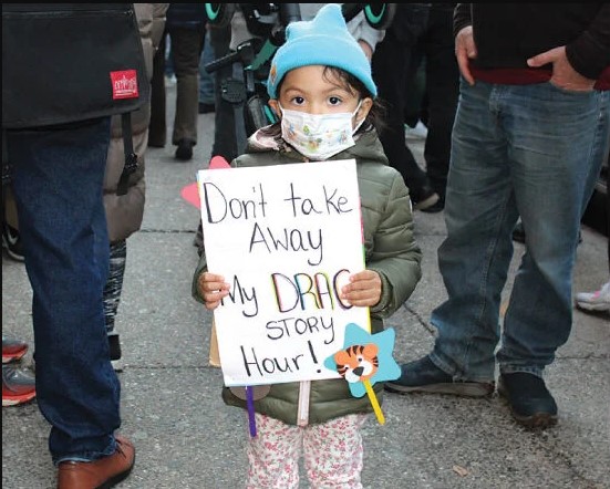 Photo of a very young child (4?) with a small sign reading "Don't take away my DRAG STORY Hour!"